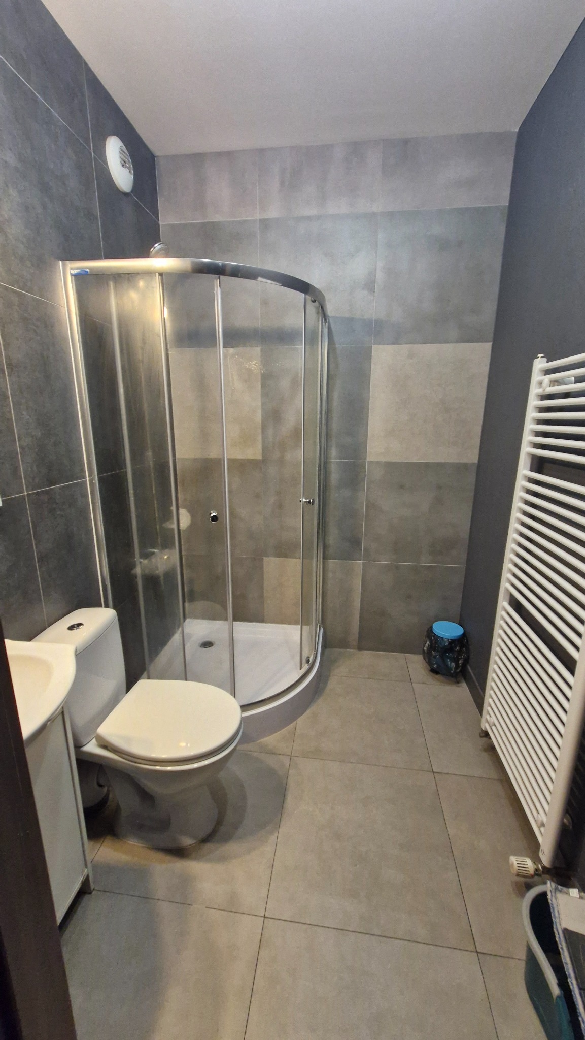 Nowoczesna łazienka z kabiną prysznicową, umywalką z szafką i grzejnikiem drabinkowym. Szaro-biała kolorystyka, płytki imitujące beton. Ukończona realizacja.