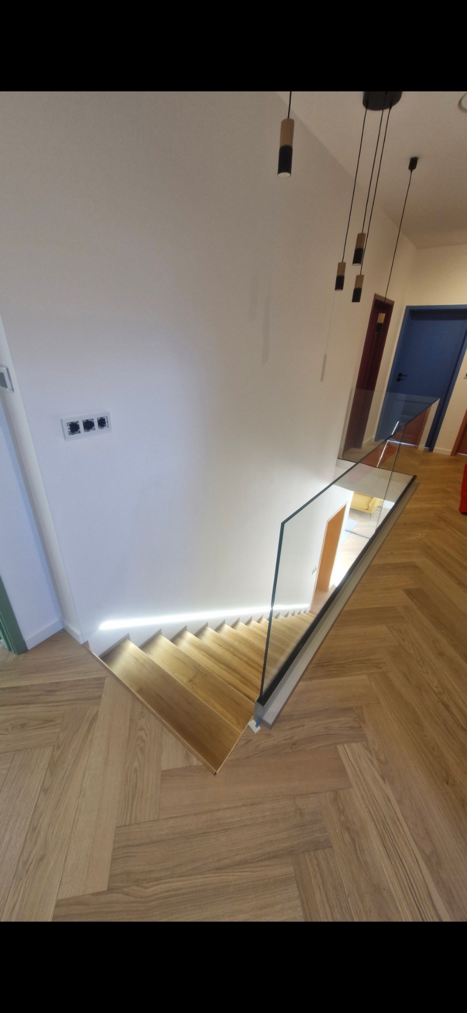 Nowoczesne schody drewniane z podświetleniem LED i szklaną balustradą, widok z góry na parkiet w jodełkę, minimalistyczne lampy wiszące.