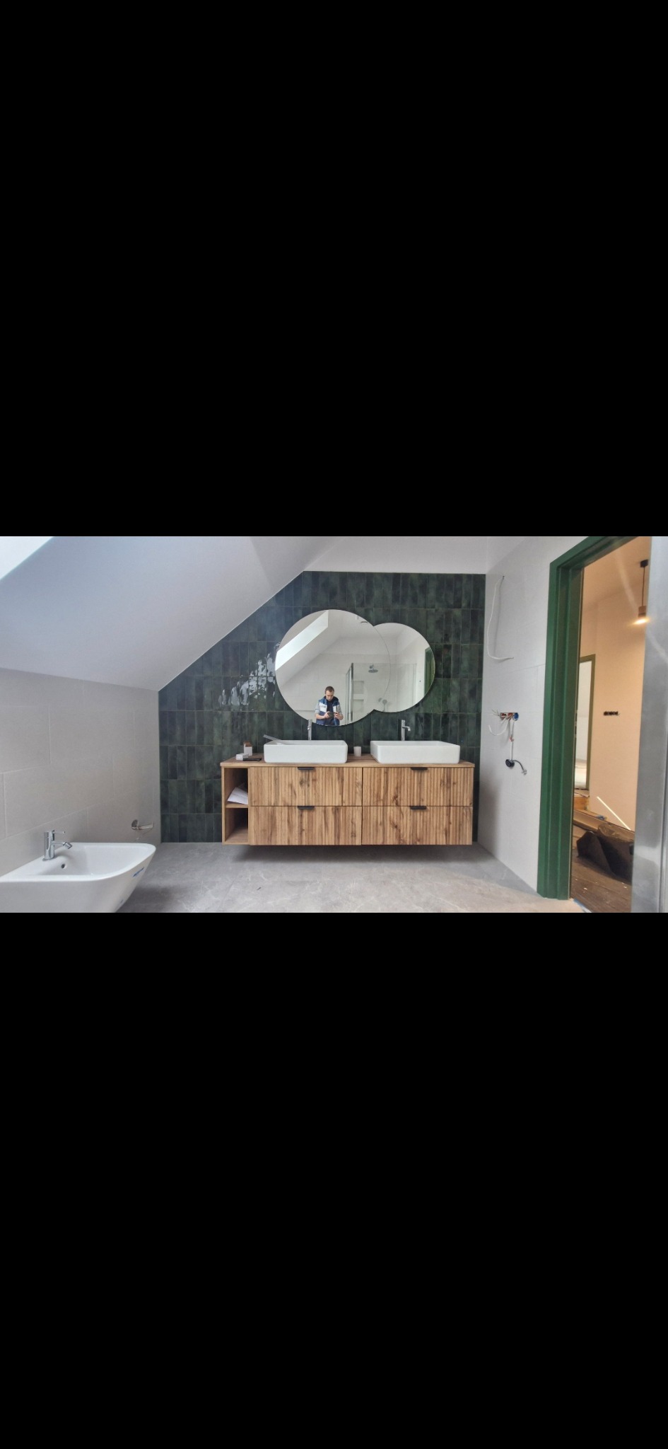 Nowoczesna łazienka na poddaszu z drewnianą szafką pod umywalki, zieloną glazurą i owalnym lustrem. W tle widoczne drzwi z zieloną ościeżnicą i bidet.