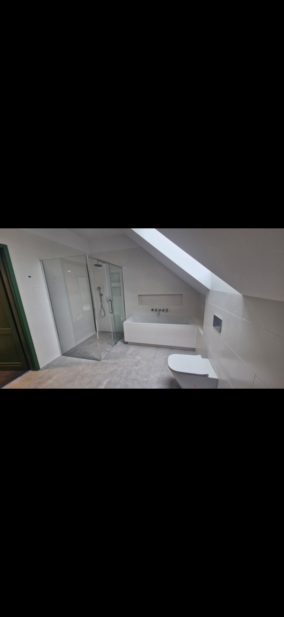 Jasna łazienka na poddaszu ze szklaną kabiną prysznicową, białą wanną i toaletą. Uwagę przykuwa okno dachowe wpuszczające naturalne światło.