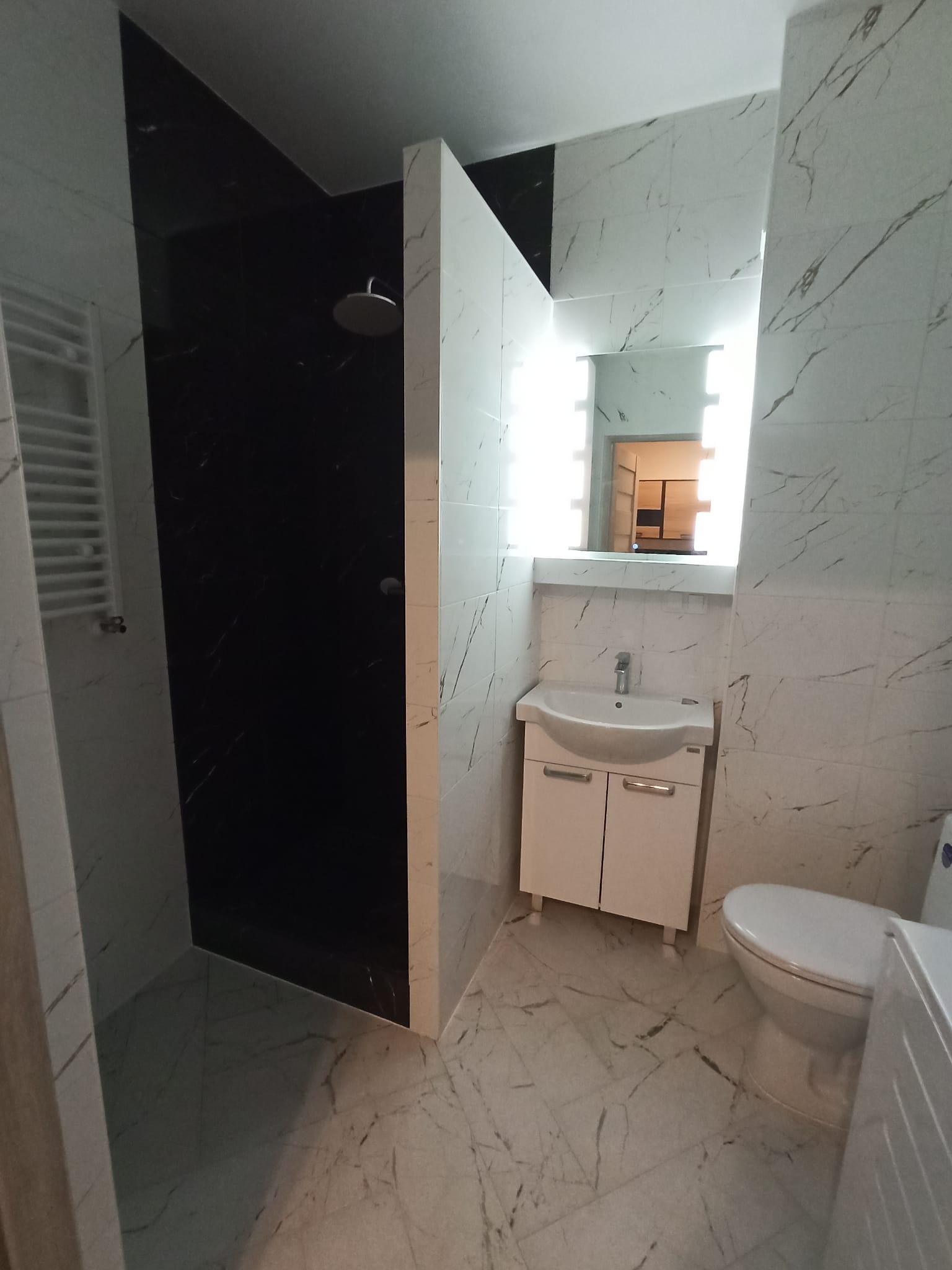 Elegancka łazienka z kabiną walk-in wykończoną czarnym marmurem, białe płytki imitujące marmur, podświetlane lustro nad umywalką i toaleta. Nowoczesny design.