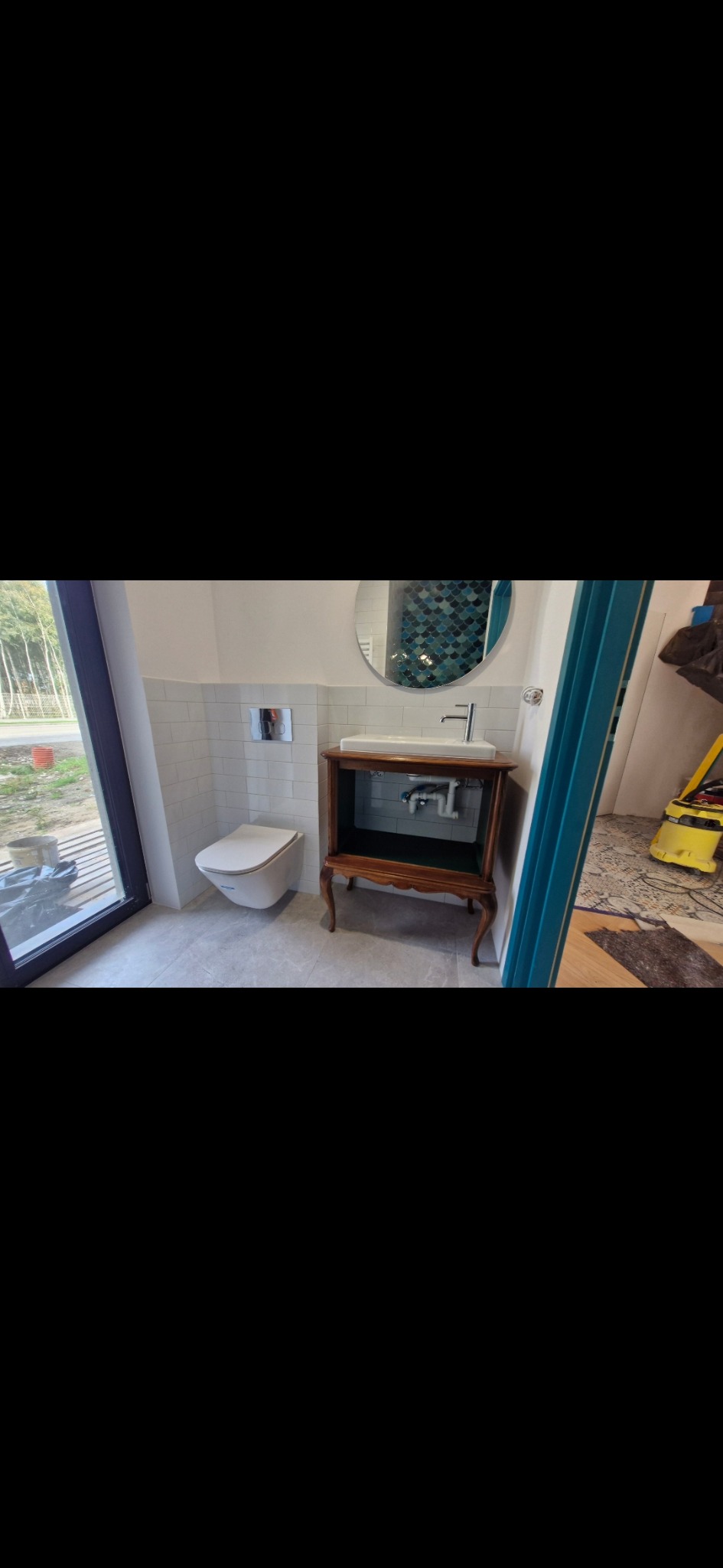 Wykończona łazienka z toaletą podwieszaną, umywalką na dekoracyjnej szafce i okrągłym lustrem z mozaiką. Widoczne płytki na ścianach i podłodze.