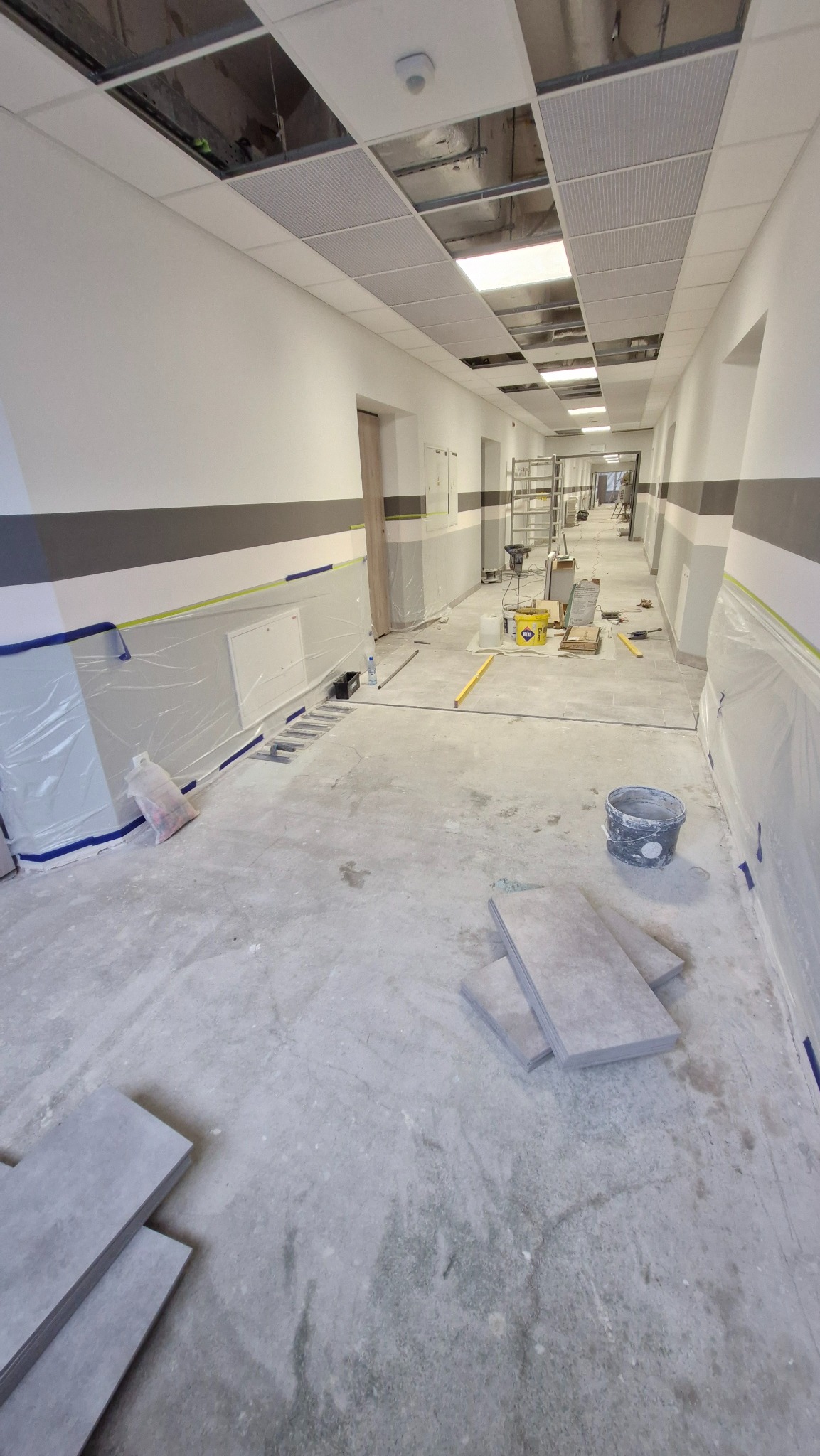 Korytarz w trakcie prac wykończeniowych: zabezpieczone ściany folią, płyty na podłodze, sufit podwieszany z otworami. Widoczne narzędzia i materiały budowlane.