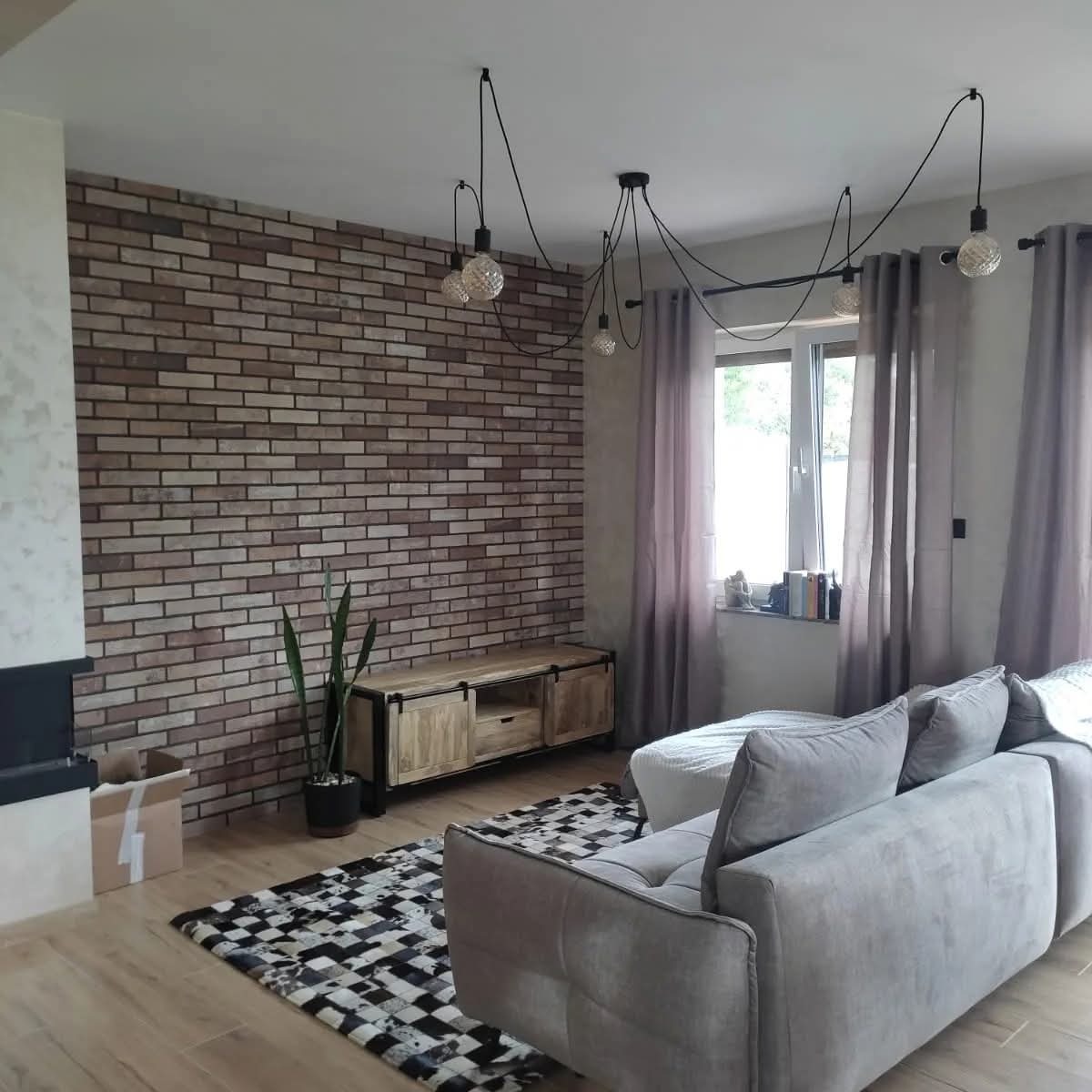 Salon z ceglaną ścianą, drewnianą komodą i designerską lampą pająk. Widoczny fragment sofy i dywan w geometryczny wzór. Nowoczesne wykończenie wnętrza.
