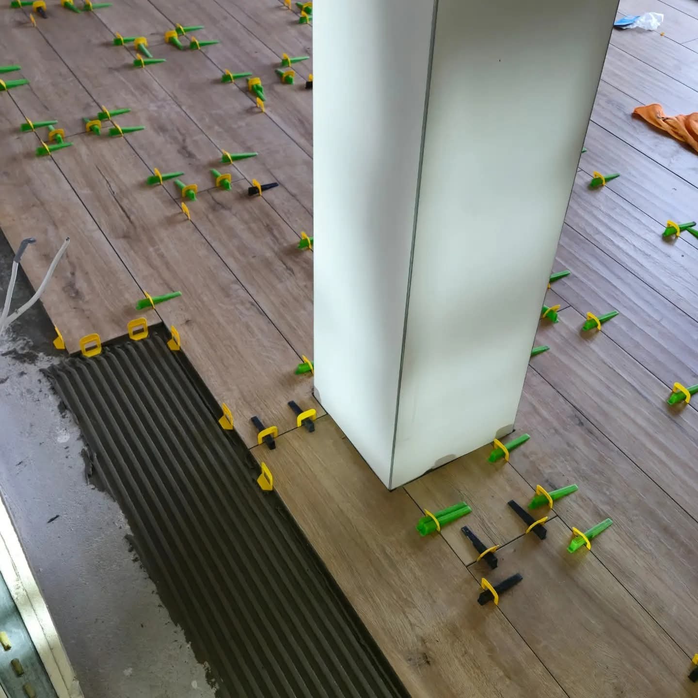 Układanie drewnopodobnych płytek podłogowych z użyciem klipsów poziomujących, widoczna warstwa kleju i fragment kolumny. Praca w toku, zbliżenie na podłogę.