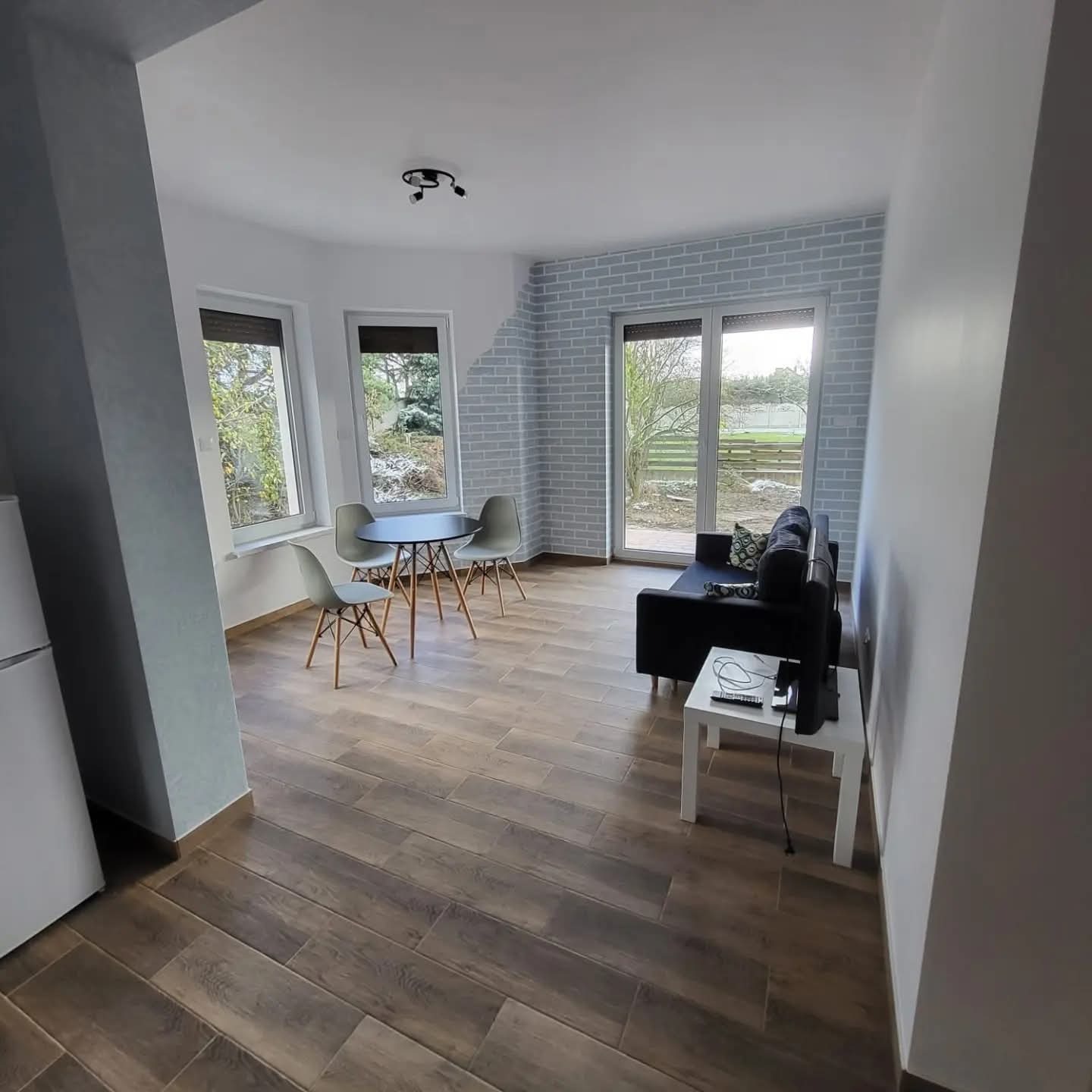 Jasne wnętrze po remoncie: drewnopodobna podłoga, ściana z efektem cegły, okna z widokiem na zieleń. Minimalistyczny stół i sofa tworzą przytulną przestrzeń.