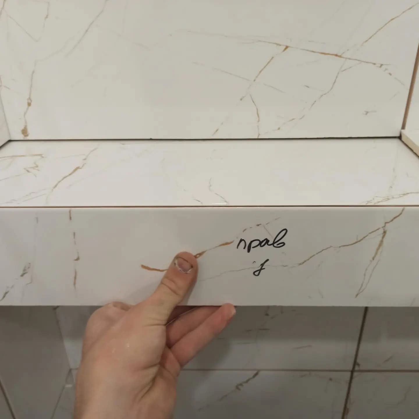 Układanie białych płytek imitujących marmur z brązowymi żyłkami. Widoczna ręka glazurnika przytrzymująca płytkę. Napis na płytce.