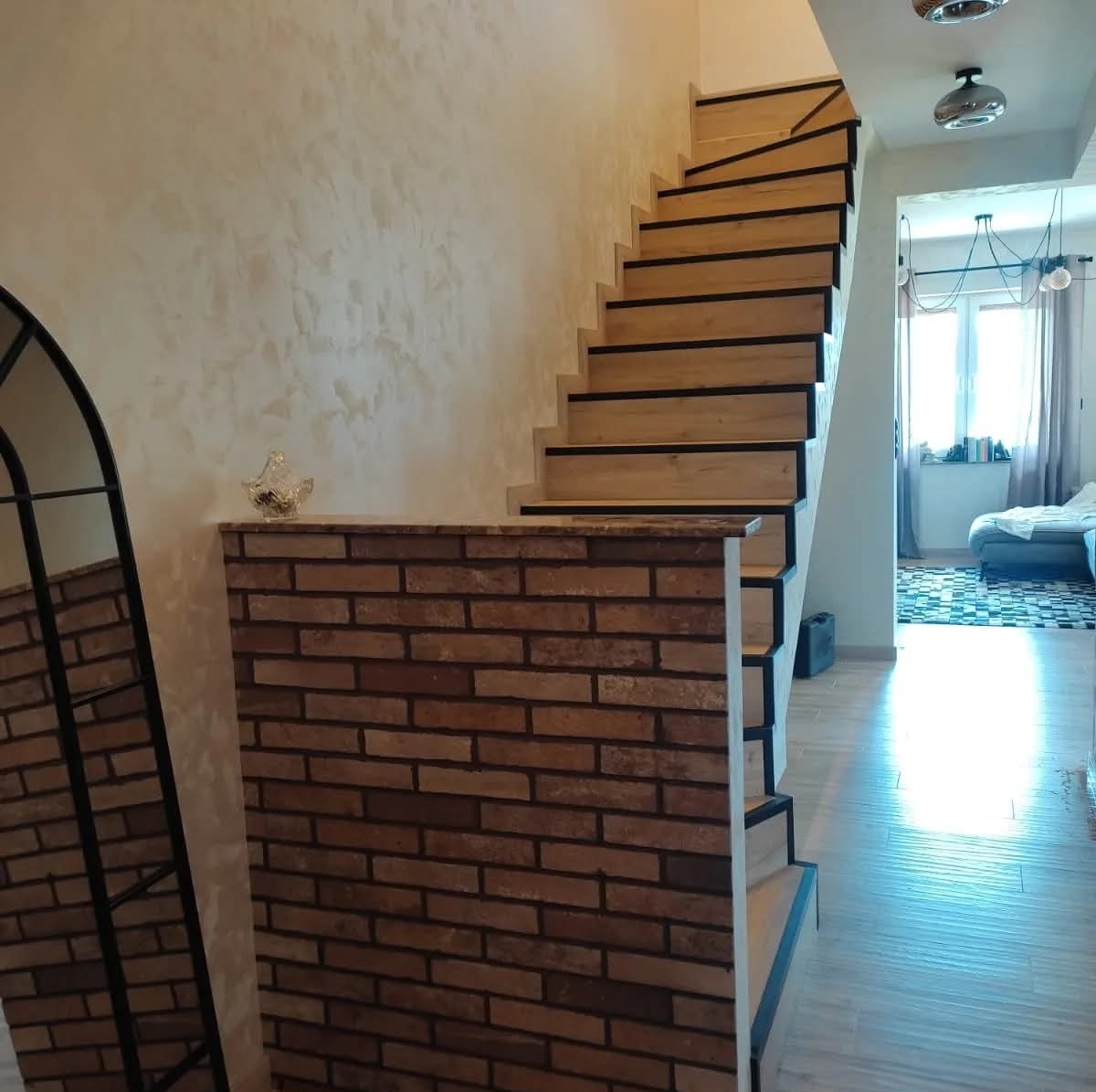 Stylowe schody z drewnianymi stopniami i czarnymi krawędziami, obok ściana z cegły i lustro. W tle widoczny salon z oknem i kanapą. Jasne, nowoczesne wnętrze.