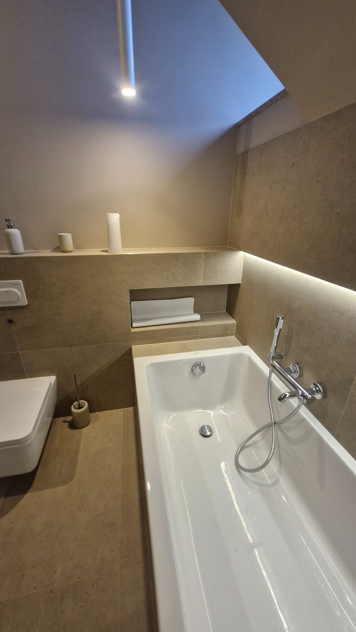 Nowoczesna łazienka w beżowych tonacjach: wanna, toaleta, półki z oświetleniem LED. Minimalistyczny design, ciepłe światło, płytki imitujące kamień.