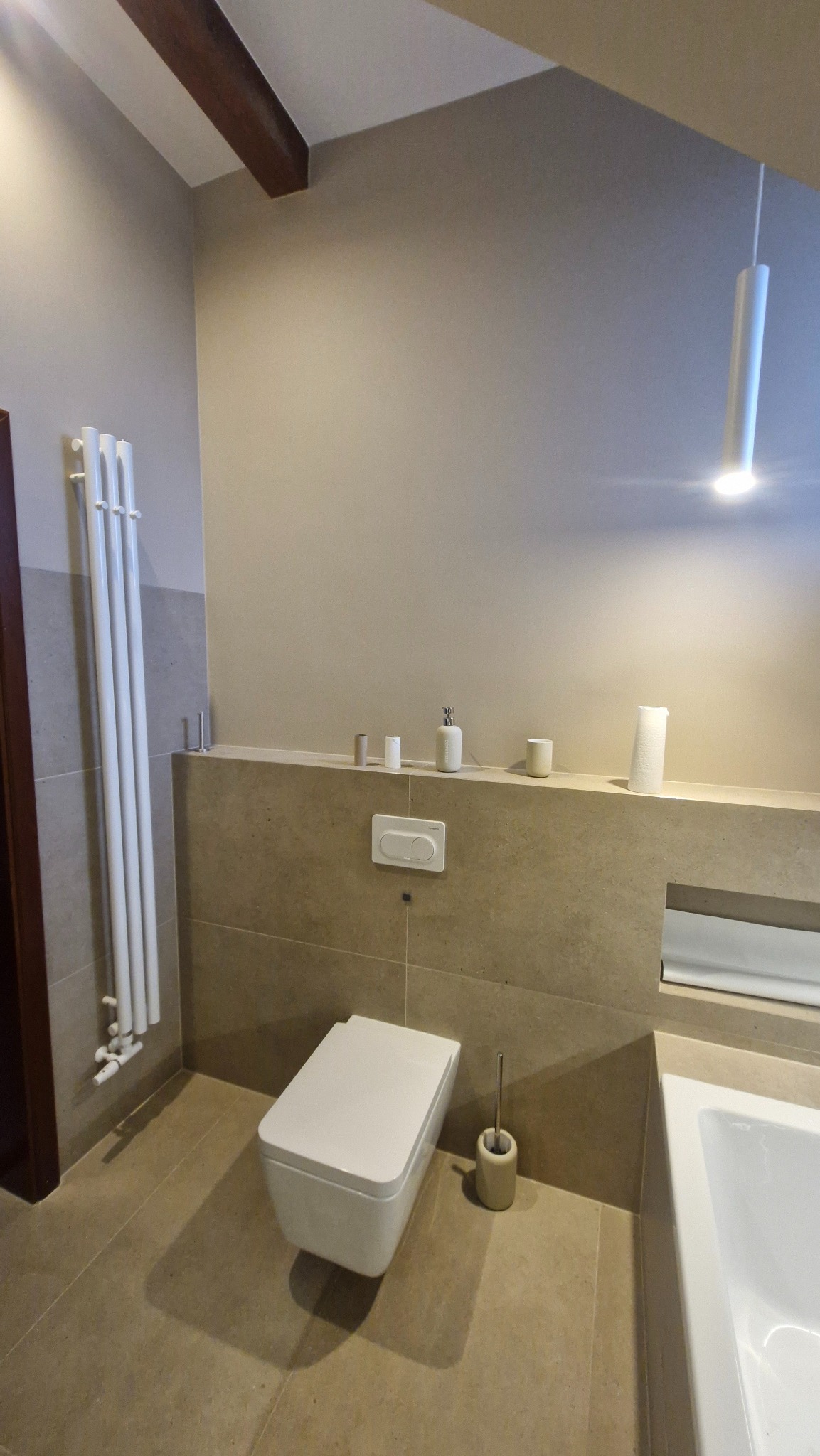 Nowoczesna łazienka z podwieszaną toaletą, beżowymi płytkami i minimalistycznym oświetleniem. Widoczny grzejnik drabinkowy i drewniany element stropu.