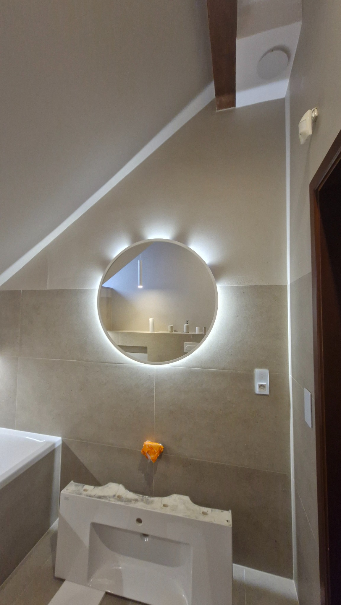 Łazienka w trakcie wykańczania: beżowe płytki, okrągłe lustro z podświetleniem LED, biała umywalka jeszcze nie zamontowana, widoczne elementy instalacji.