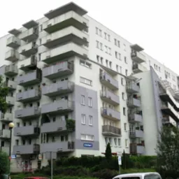 Wielopiętrowy budynek mieszkalny z balkonami w szaro-białej kolorystyce, widoczny z poziomu ulicy z elementami zieleni i zaparkowanymi samochodami.