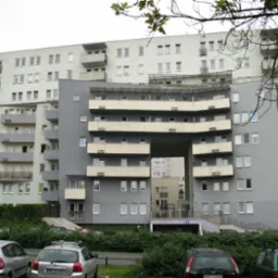 Wielopiętrowy budynek mieszkalny z żelbetową konstrukcją, widoczne balkony i przejście w bryle budynku, zieleń i zaparkowane samochody przed budynkiem.