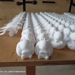 Rząd białych, małych figurek kaczek z misiami i dinozaurów, wykonanych metodą addytywną, ułożonych na drewnianej powierzchni. Duża ilość detali, jasne oświetlenie.