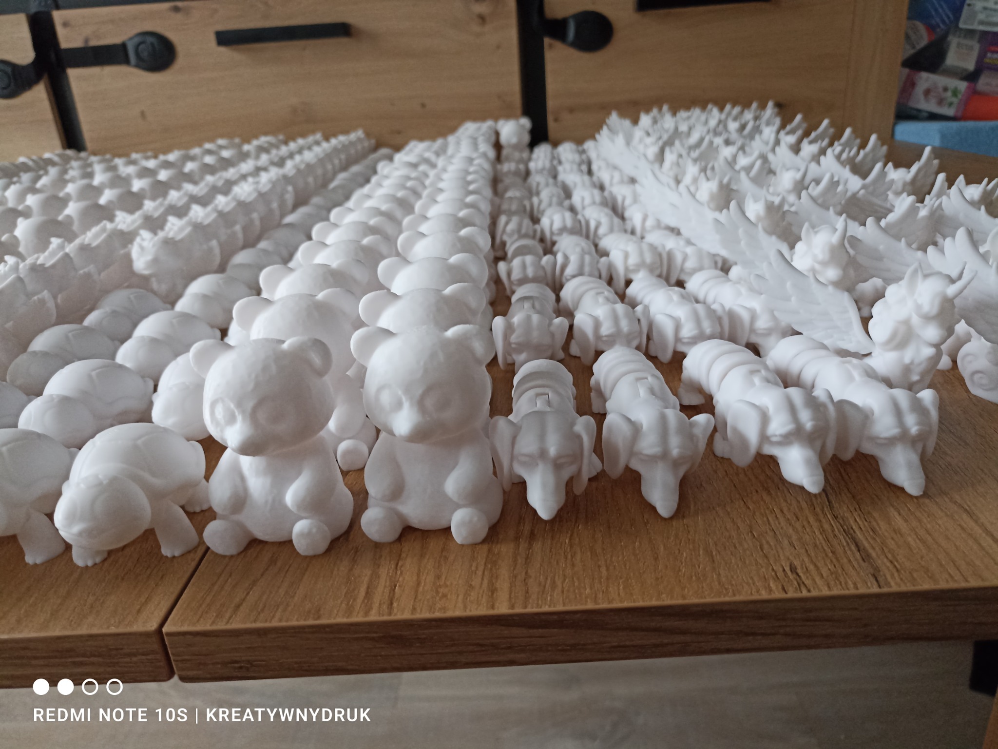 Seria białych figurek: misie, żółwie, psy i skrzydlate stworzenia, ustawione w rzędach na drewnianej powierzchni. Wyraźne detale i tekstura.