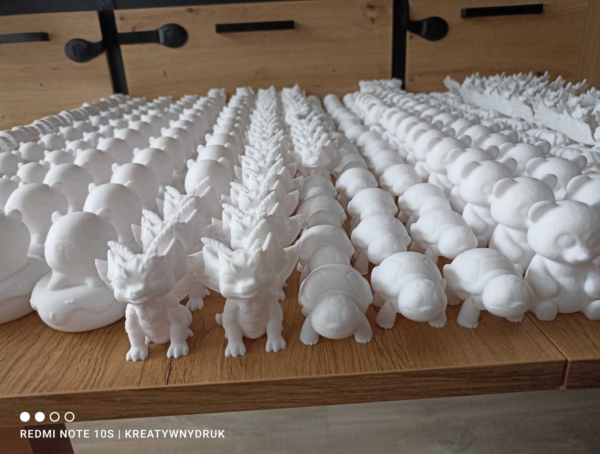 Rządki białych, małych figurek: smok, żółw, miś, kaczka. Figurki ustawione na drewnianej powierzchni. Widoczny napis 'KREATYWNYDRUK'.