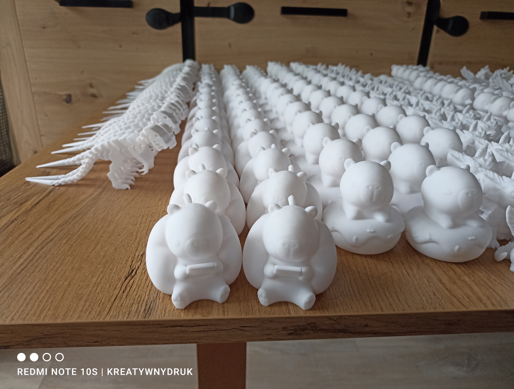 Rząd białych, małych figurek kaczek z misiami i dinozaurów, wykonanych metodą addytywną, ułożonych na drewnianej powierzchni. Duża ilość detali, jasne oświetlenie.