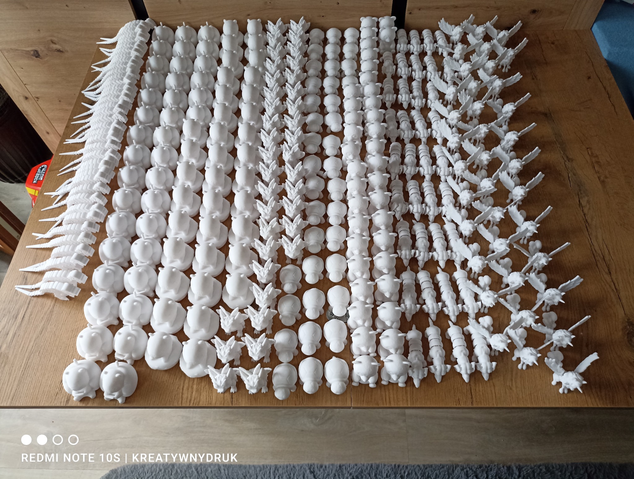 Różnorodne, białe elementy wykonane metodą addytywną, ułożone rzędami na drewnianym stole. Widoczne detale modeli, od prostych kształtów po skomplikowane figurki.