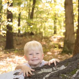 Klaudia Boguszewska - Uśmiechnięty blond chłopiec, oparty o spróchniały pień drzewa w lesie, spogląda w obiektyw. Część pnia pokryta mchem. W tle drzewa i liście.