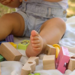 Klaudia Boguszewska - Dziecięca stopa wśród drewnianych klocków na białym kocyku, w tle zieleń. Niska perspektywa, miękkie światło, naturalna kolorystyka. Ujęcie z sesji rodzinnej.