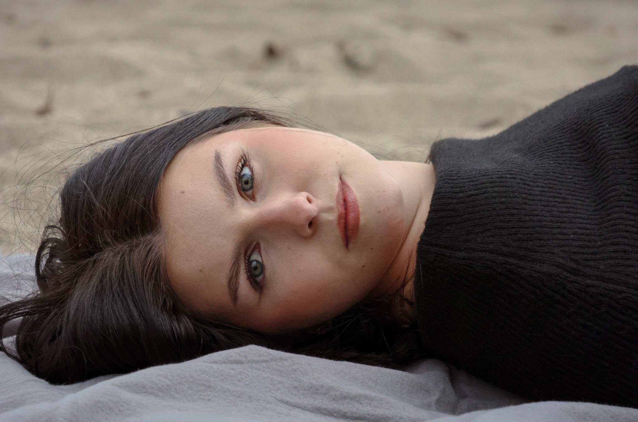 Portret młodej kobiety z ciemnymi włosami i zielonymi oczami, leżącej na jasnym materiale, z piaskiem w tle. Delikatne oświetlenie podkreśla naturalne piękno.