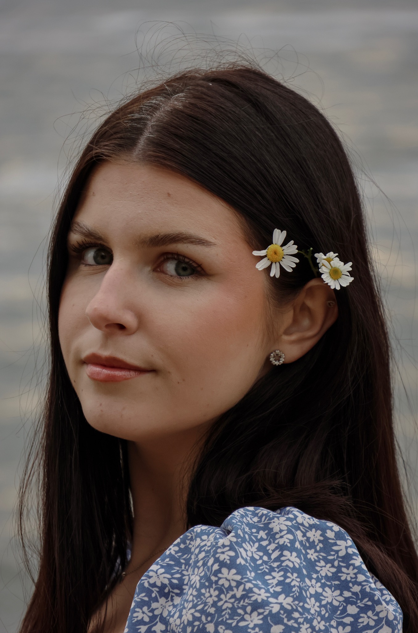 Portret kobiety z ciemnymi włosami, ozdobionymi stokrotkami za uchem, w niebieskiej sukience w kwiaty, spoglądającej w bok na neutralnym tle.