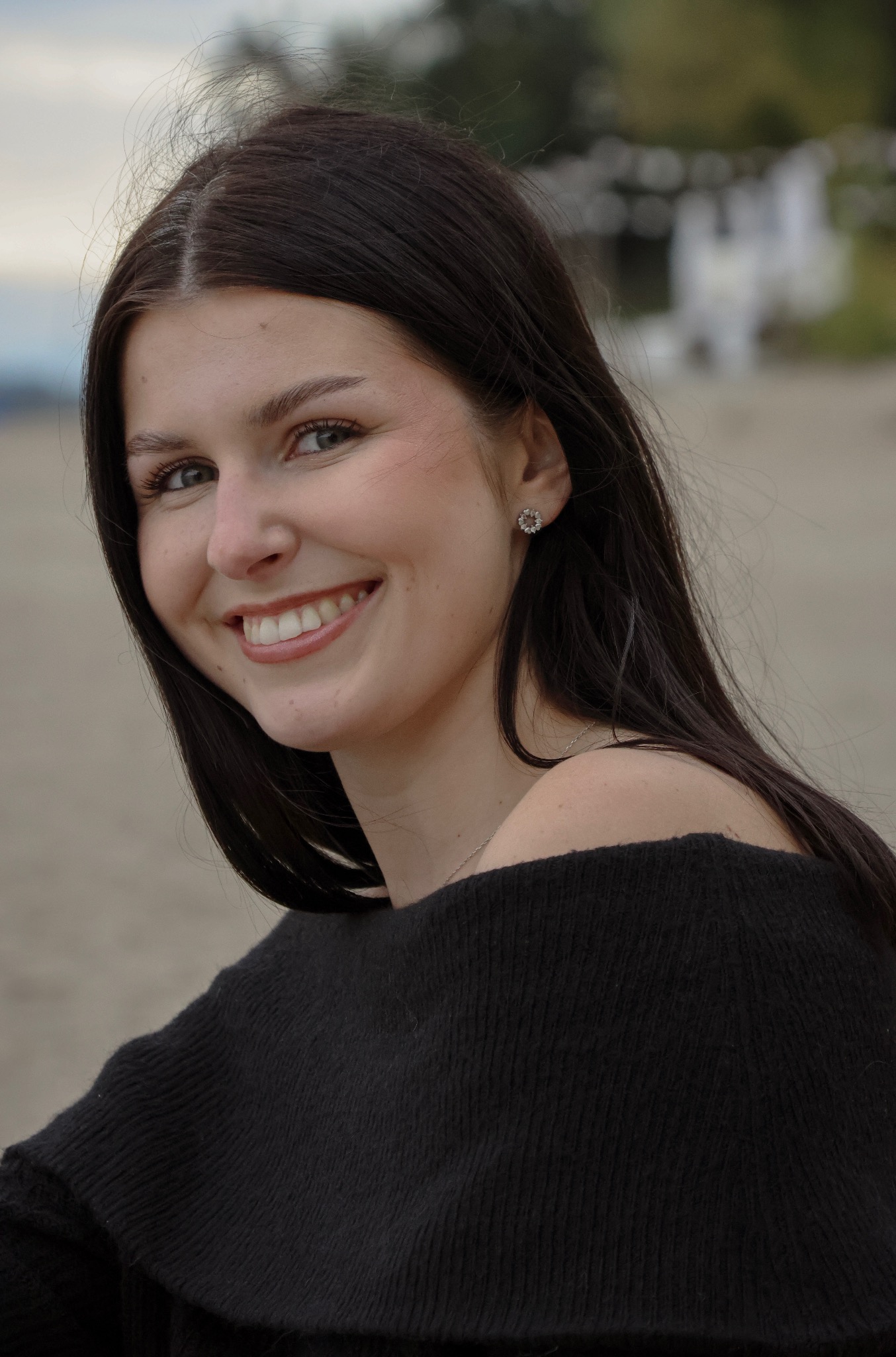 Portret uśmiechniętej brunetki w czarnym swetrze na tle plaży. Widoczne kolczyki i delikatny łańcuszek. Naturalne światło podkreśla blask uśmiechu.