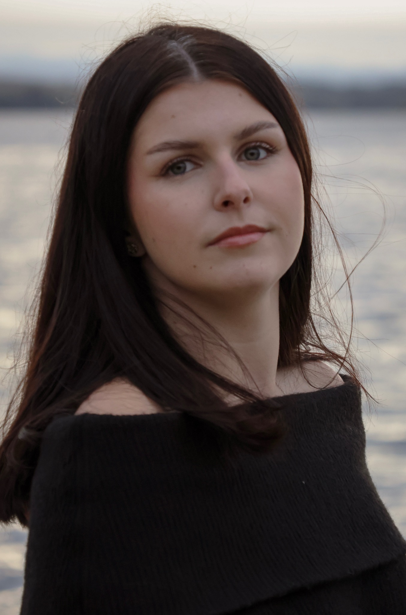 Portret młodej kobiety z ciemnymi włosami, na tle wody i zachmurzonego nieba, w czarnym swetrze z odkrytymi ramionami. Delikatny makijaż, wiatr we włosach.