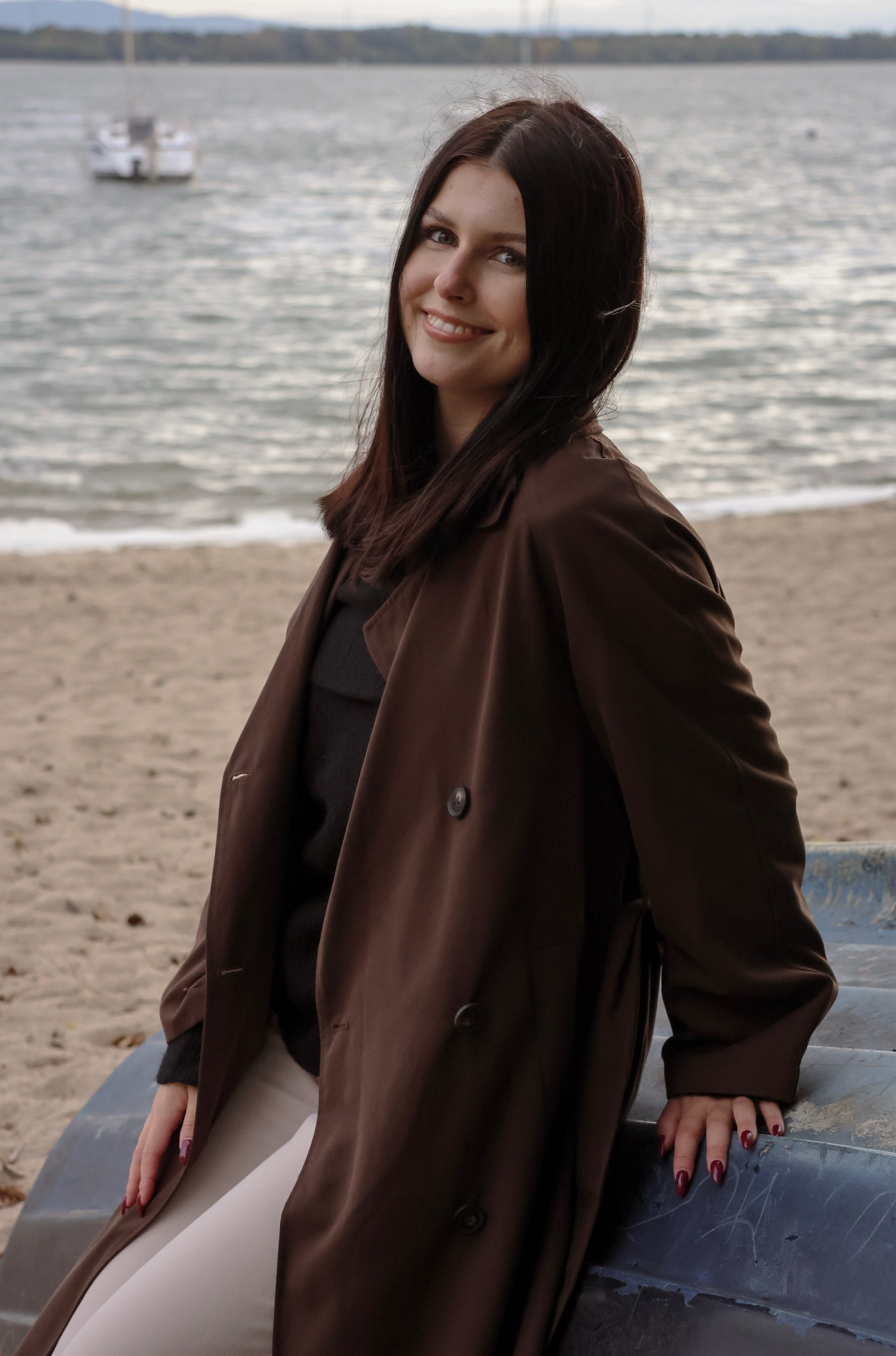 Uśmiechnięta brunetka w brązowym płaszczu na tle plaży i morza, opierająca się o łódź, z widocznym jachtem w oddali. Portret w naturalnym świetle.