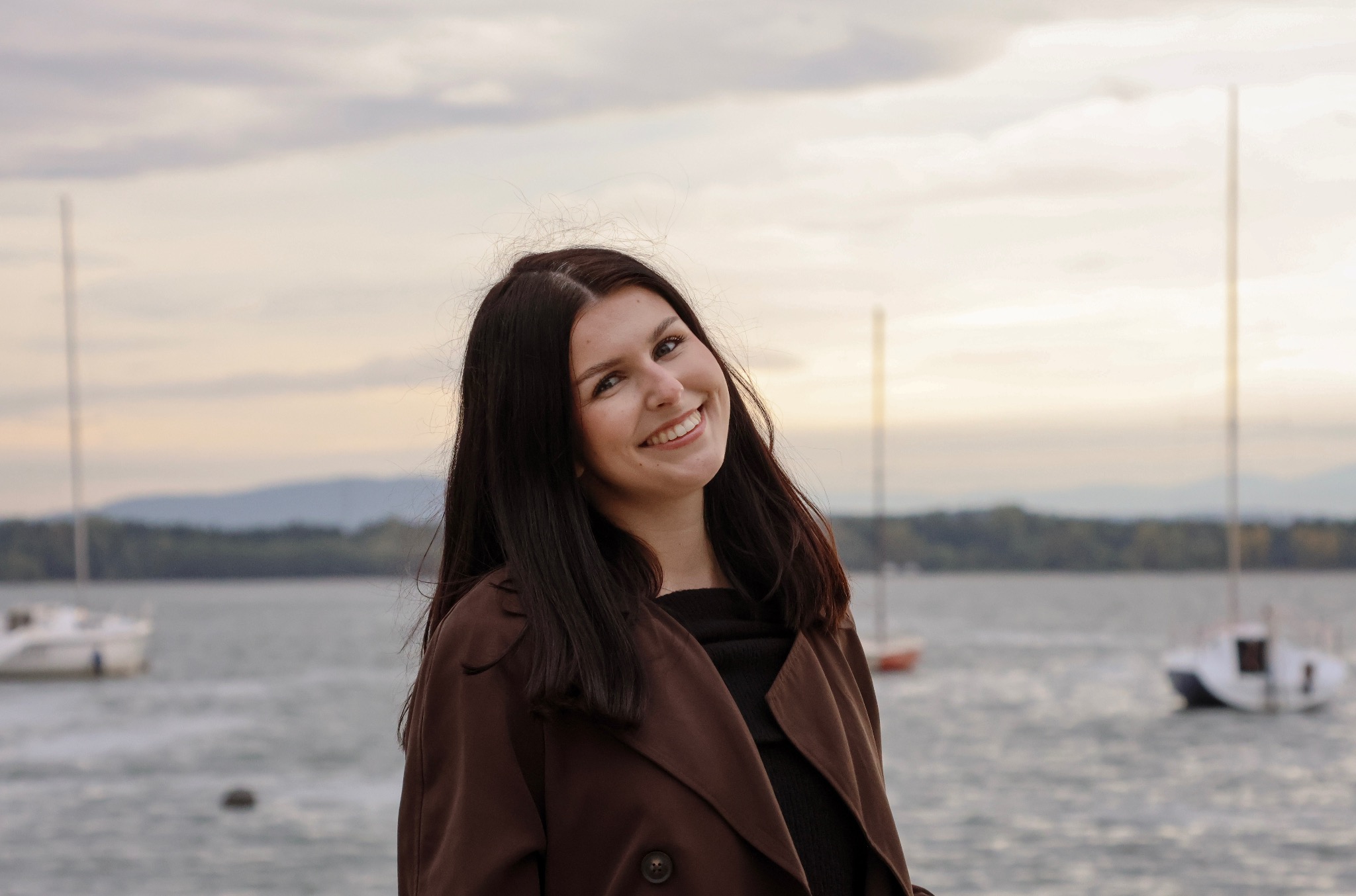 Uśmiechnięta brunetka w brązowym płaszczu na tle jeziora z łódkami. Delikatne światło, naturalny portret.