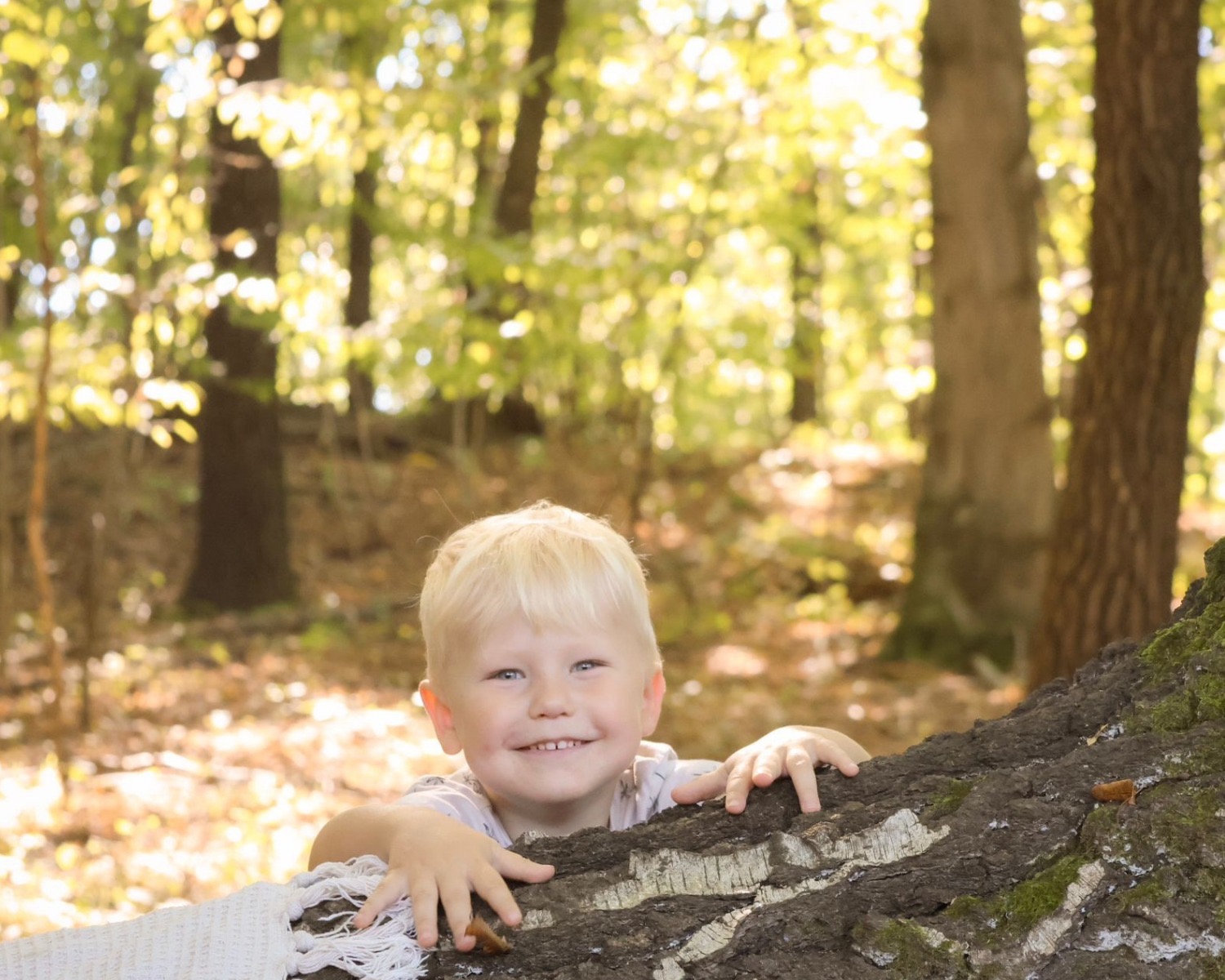 Uśmiechnięty blond chłopiec, oparty o spróchniały pień drzewa w lesie, spogląda w obiektyw. Część pnia pokryta mchem. W tle drzewa i liście.