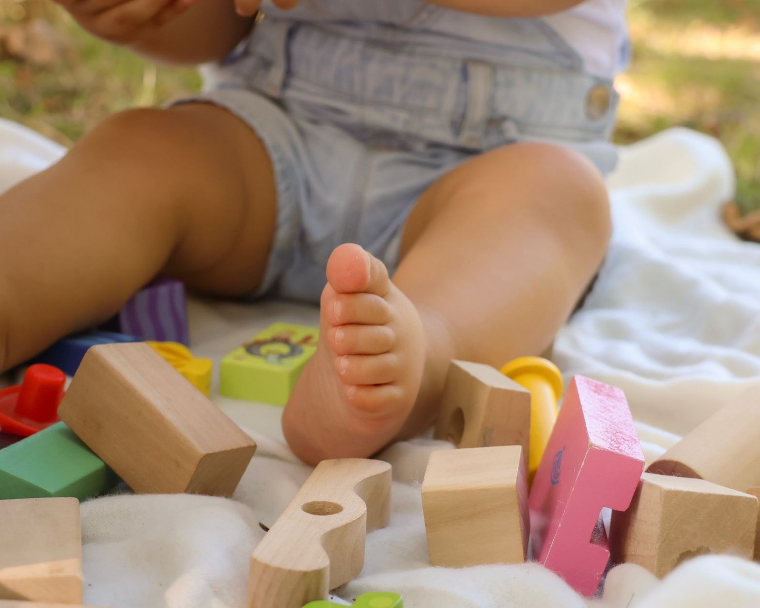 Dziecięca stopa wśród drewnianych klocków na białym kocyku, w tle zieleń. Niska perspektywa, miękkie światło, naturalna kolorystyka. Ujęcie z sesji rodzinnej.