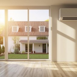 GrantEko Janina Kucińska - Klimatyzator ścienny w jasnym wnętrzu z dużym oknem na dom z ogrodem. Parkiet odbija promienie słoneczne. Roślina doniczkowa w lewym rogu.