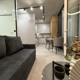 Renov8 Complex Renovation - Nowoczesne wnętrze mieszkania po remoncie: szary narożnik, okrągły stolik kawowy, otwarta kuchnia z białymi szafkami i drewnianym akcentem. Oświetlenie punktowe.