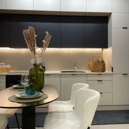 Renov8 Complex Renovation - Nowoczesna kuchnia w bieli i czerni z drewnianym koszykiem na blacie i dekoracyjnym wazonem na stole. Minimalistyczny design, oświetlenie LED pod szafkami.