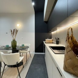 Renov8 Complex Renovation - Nowoczesna kuchnia z czarnym stołem i białymi krzesłami. Minimalistyczne szafki kuchenne z oświetleniem LED. Wazon z trawami na stole. Koszyk na blacie.
