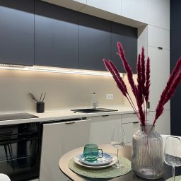 Renov8 Complex Renovation - Nowoczesna kuchnia po remoncie: minimalistyczne szafki w odcieniach szarości i bieli, oświetlenie LED podszafkowe, stół z dekoracjami.