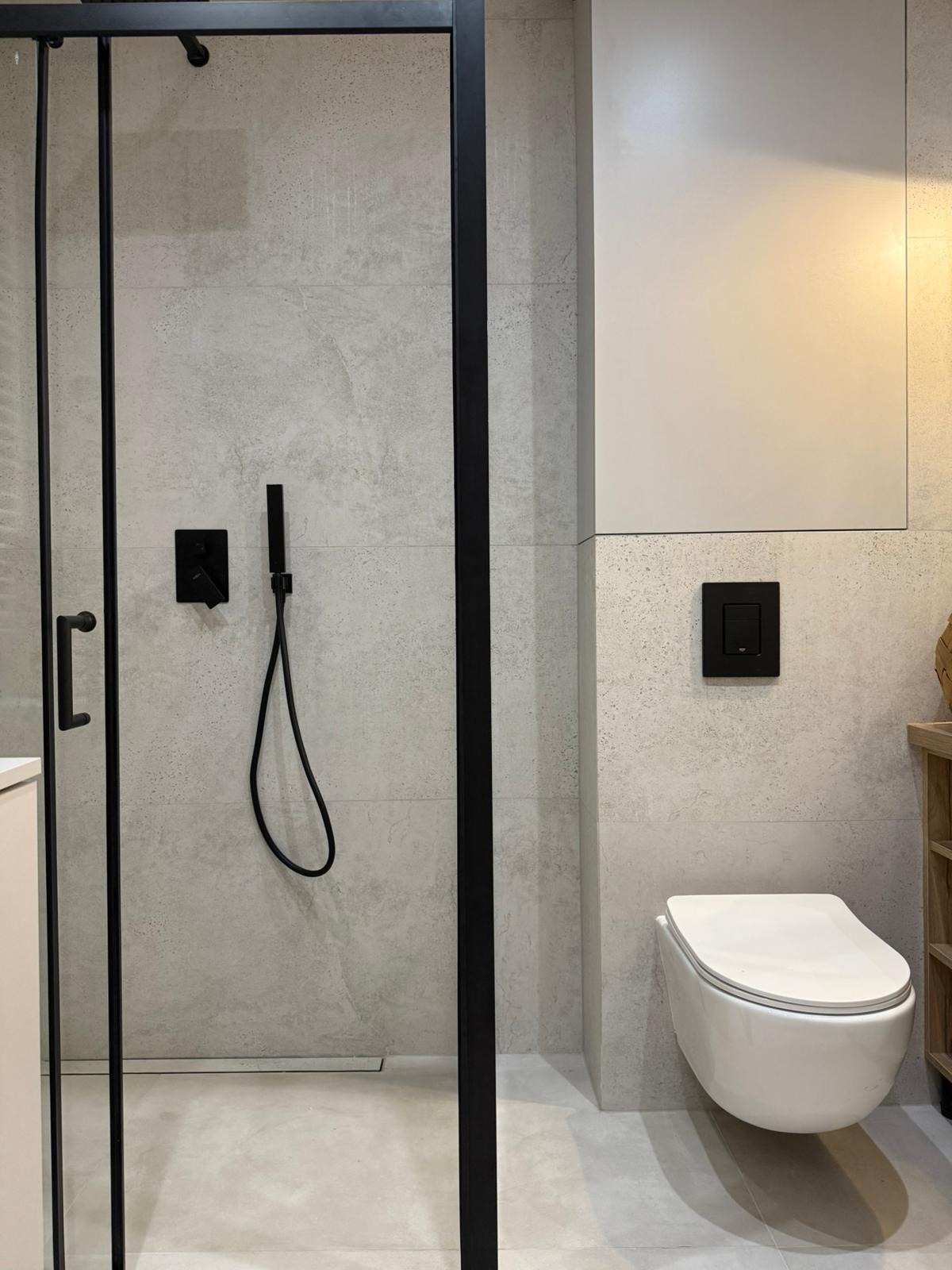 Nowoczesna łazienka po remoncie: kabina prysznicowa typu walk-in z czarnymi ramami, podwieszana toaleta i płytki imitujące beton. Minimalistyczny design, stonowana kolorystyka.