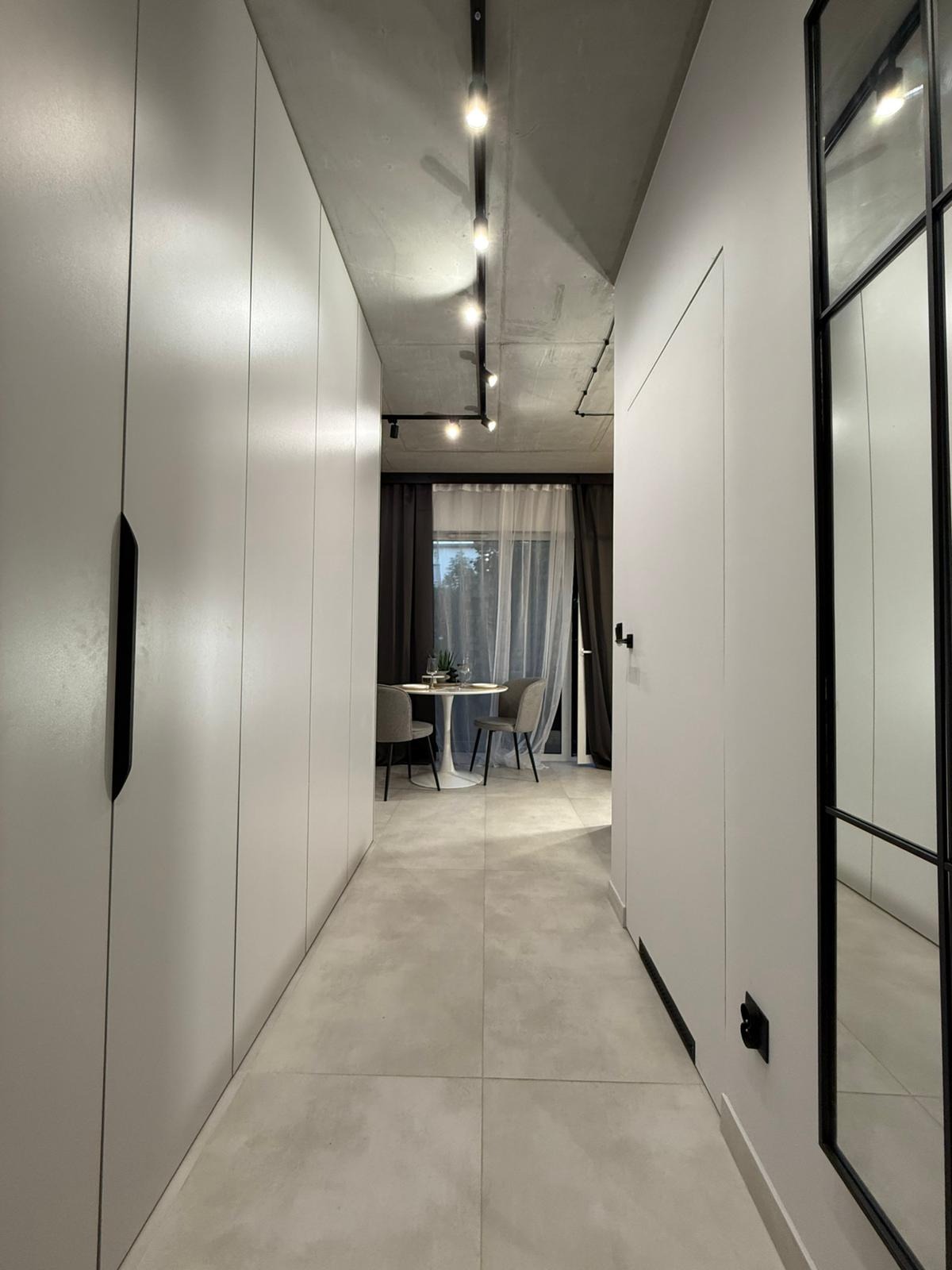 Wnętrze mieszkania w jasnych barwach. Długi korytarz z zabudową szaf w kolorze złamanej bieli prowadzi do jadalni z okrągłym stołem i krzesłami. Widoczne oświetlenie szynowe.