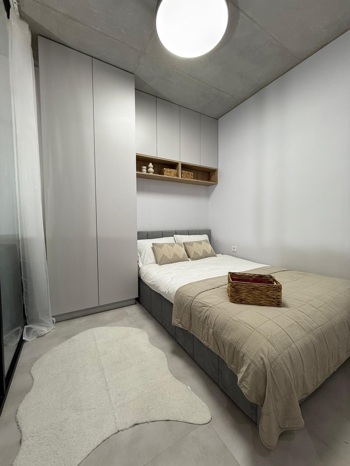 Nowoczesna sypialnia w stonowanej kolorystyce: szare ściany, betonowy sufit, zabudowa meblowa i łóżko z narzutą. Uzupełnieniem jest puszysty dywan.