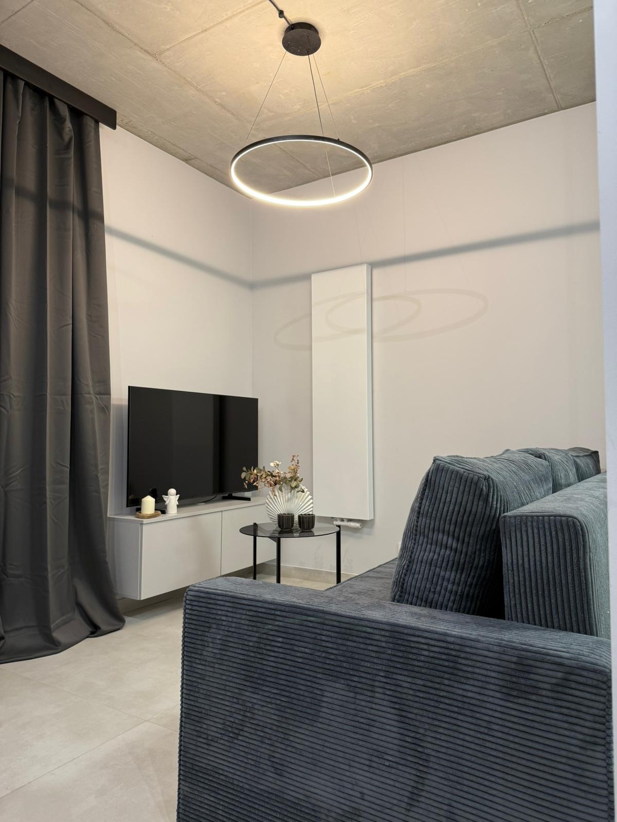 Nowoczesny salon po remoncie: szara sofa z weluru, okrągła lampa LED, telewizor na szafce RTV. Minimalistyczny design, neutralna kolorystyka, betonowy sufit.