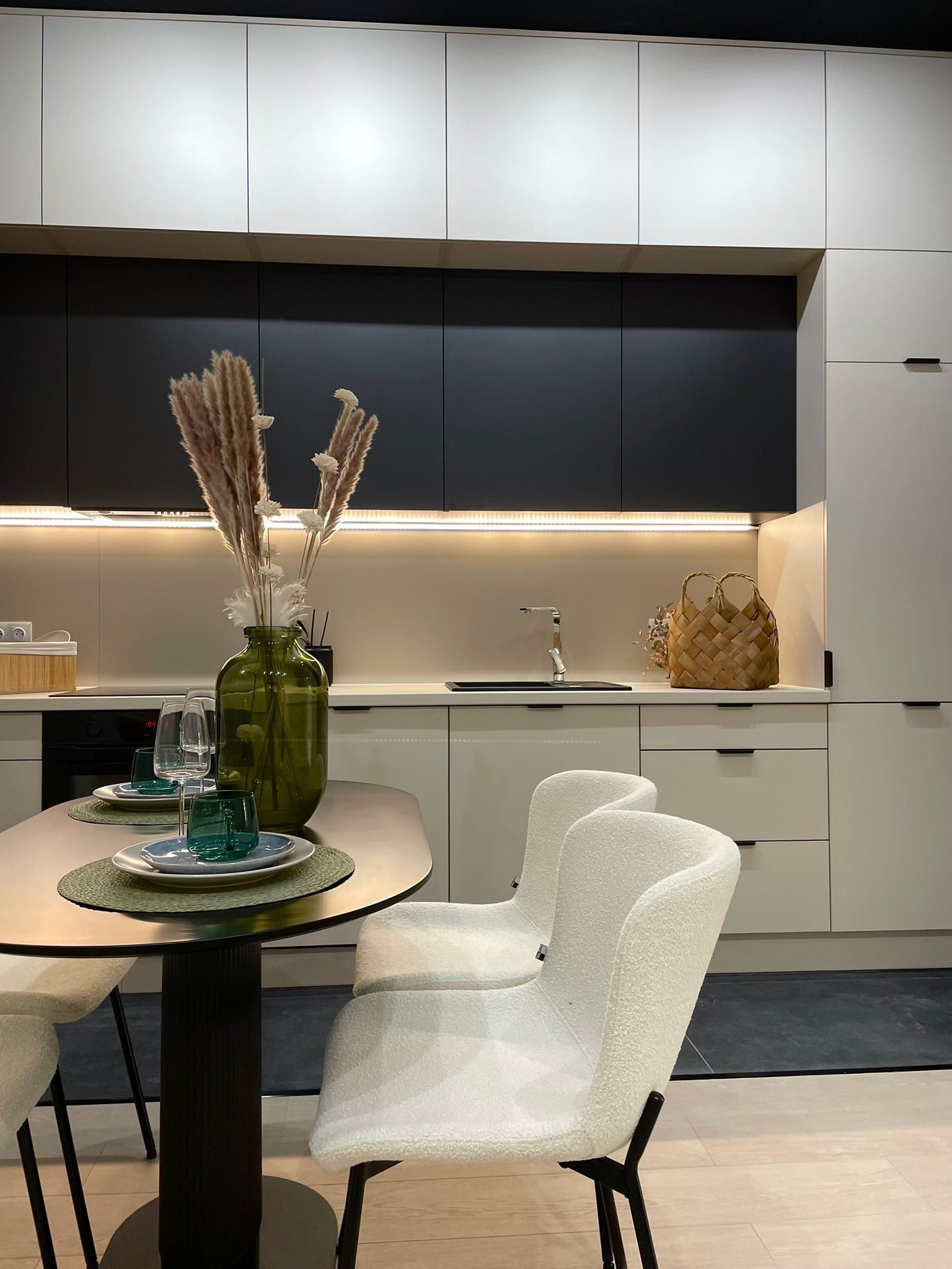 Nowoczesna kuchnia w bieli i czerni z drewnianym koszykiem na blacie i dekoracyjnym wazonem na stole. Minimalistyczny design, oświetlenie LED pod szafkami.