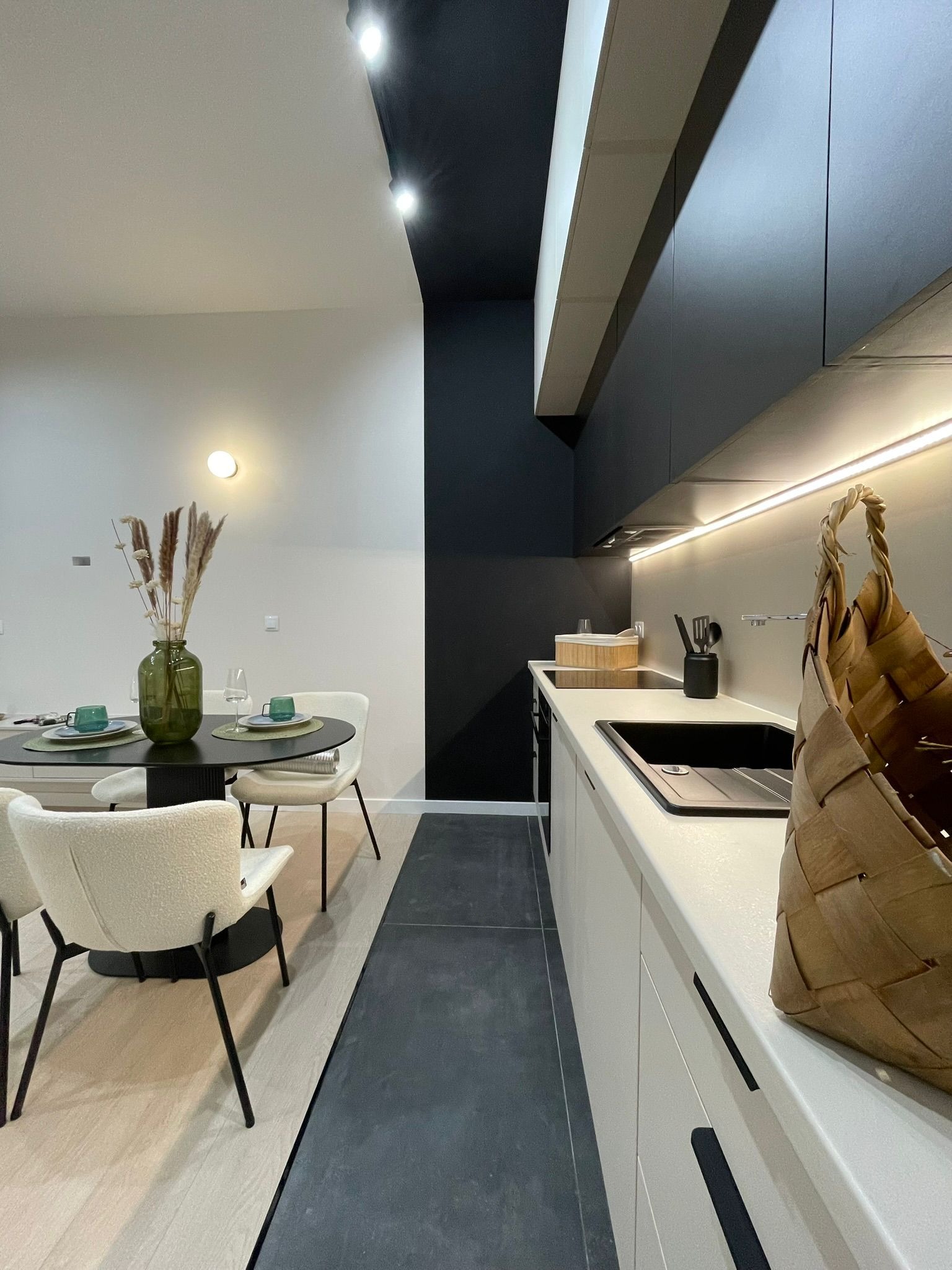 Nowoczesna kuchnia z czarnym stołem i białymi krzesłami. Minimalistyczne szafki kuchenne z oświetleniem LED. Wazon z trawami na stole. Koszyk na blacie.