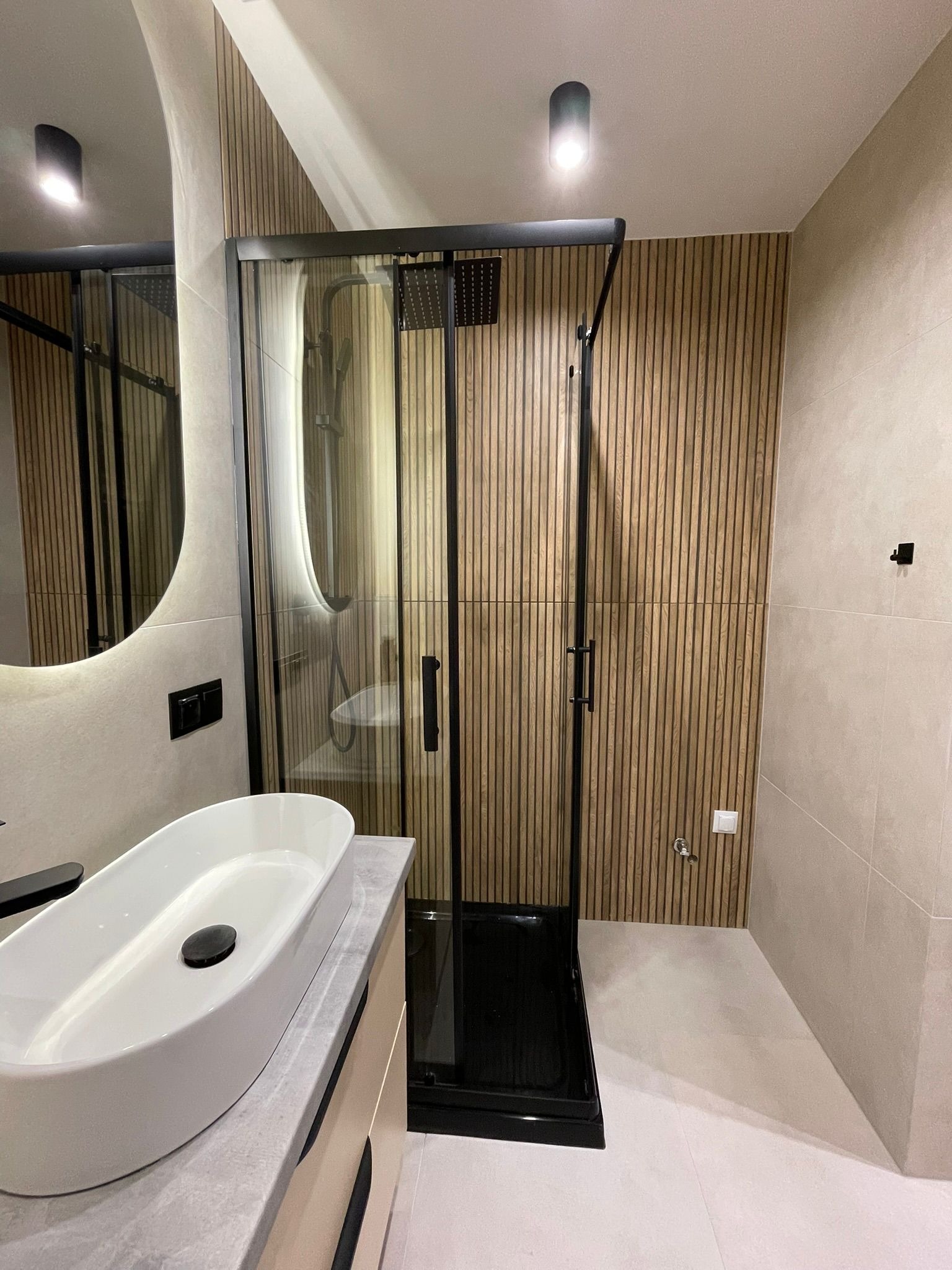 Nowoczesna łazienka z kabiną prysznicową w czarnej ramie, umywalką nablatową i lustrem z podświetleniem. Ściana wykończona drewnianymi panelami.
