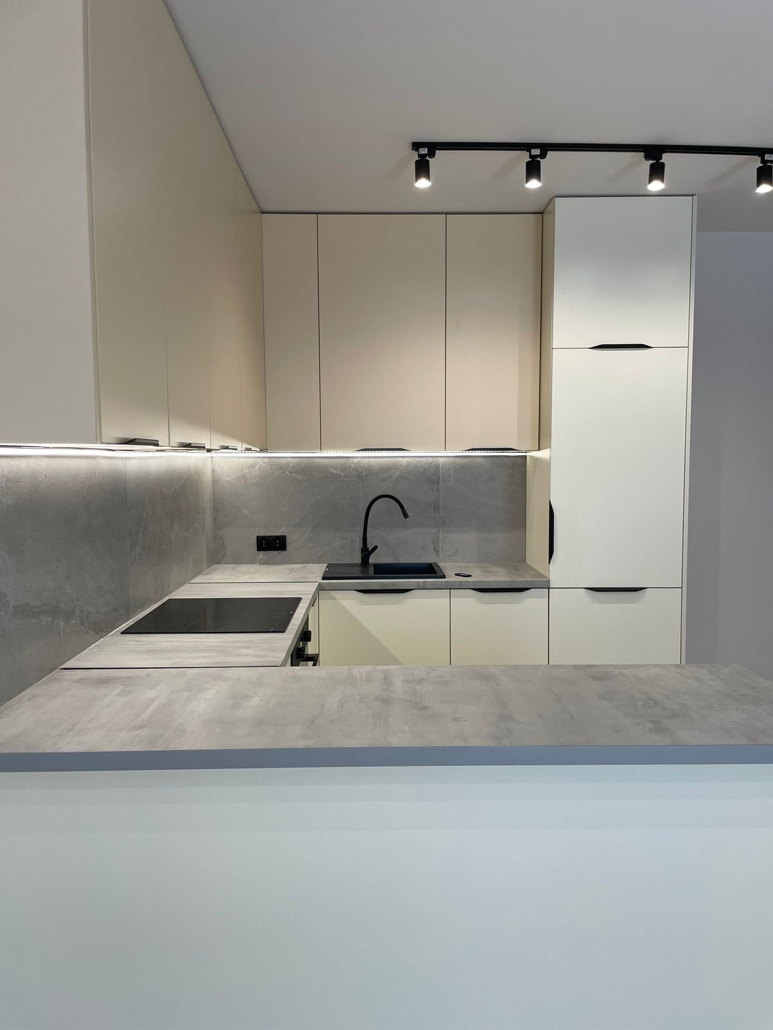 Jasna, nowoczesna kuchnia z szarym blatem i czarną armaturą. Oświetlenie podszafkowe LED i reflektory sufitowe. Minimalistyczny design i funkcjonalność.