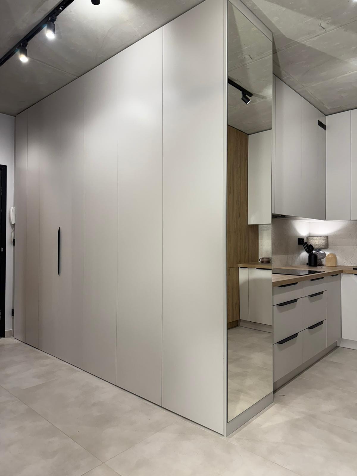 Nowoczesna kuchnia z zabudową meblową w odcieniach szarości, z lustrzaną ścianą. Widoczny fragment blatu i szafek. Minimalistyczny design, surowy beton na suficie.