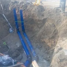 Wykop z dwoma niebieskimi rurami kanalizacyjnymi, połączonymi zielonym sznurkiem, widoczne narzędzia i fragment osoby w tle. Wykonane przyłącze wod-kan.