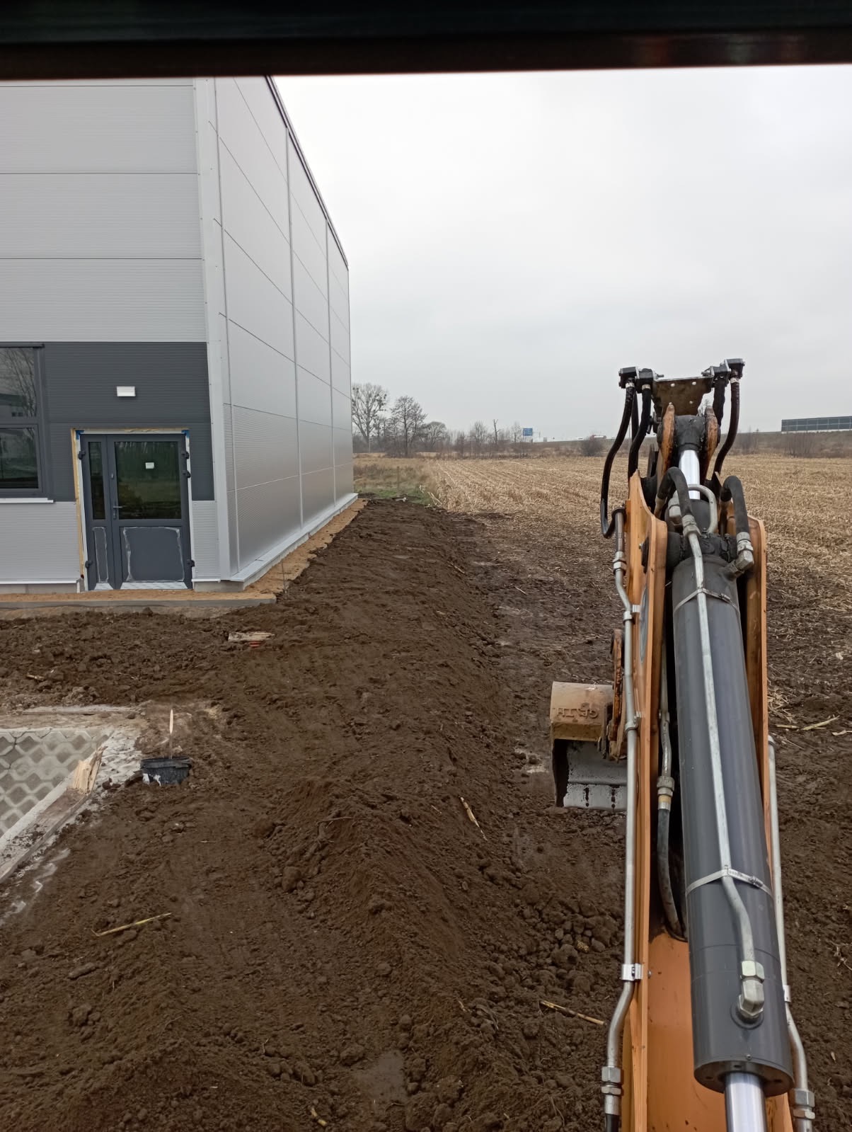 Pomarańczowa koparka w trakcie wykopu ziemi obok budynku o szarej elewacji, na tle pola i pochmurnego nieba. Widok z kabiny operatora.