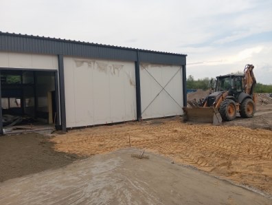 Żółty traktor przy nowym budynku z szarego metalu i białej płyty warstwowej. Przygotowanie terenu pod budowę, widoczne ślady opon i piasek.