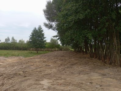 Wyrównany teren pod budowę z rzędem drzew po prawej stronie. Ślady opon na ziemi sugerują użycie ciężkiego sprzętu. W tle łąka i drzewa.